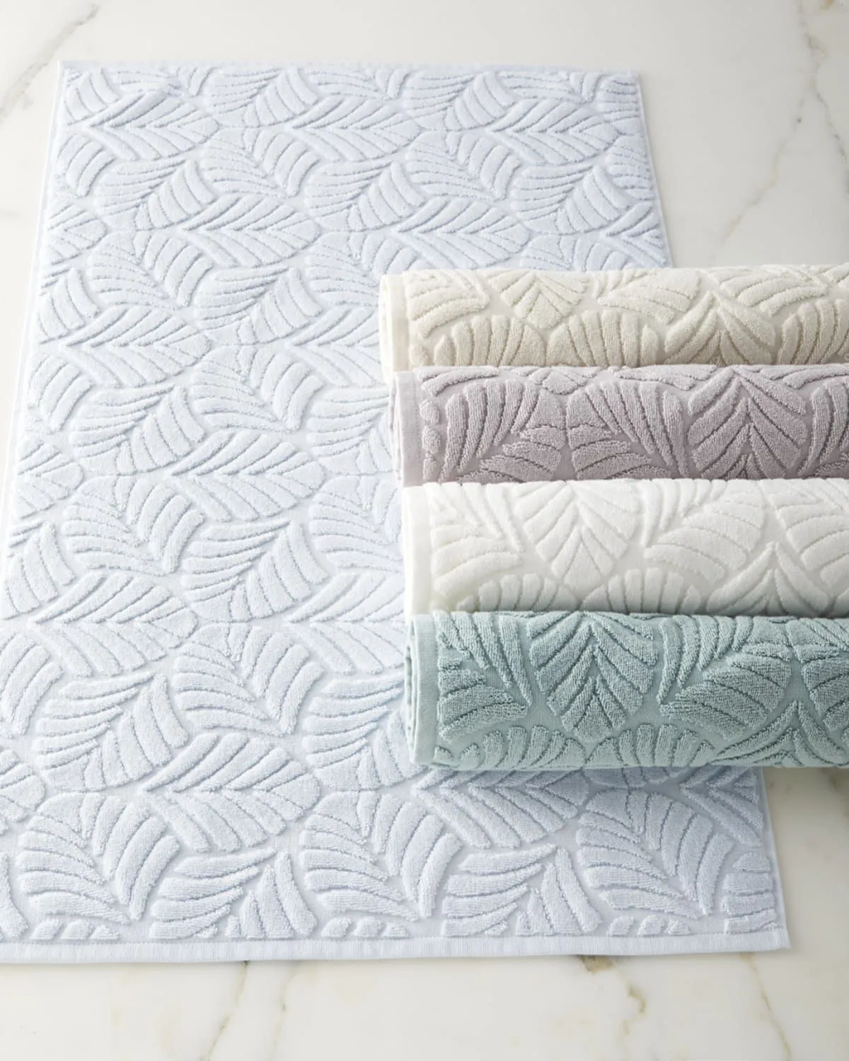 White jacquard bath mat with a leaf pattern and folded towels in cream, lilac and sage
